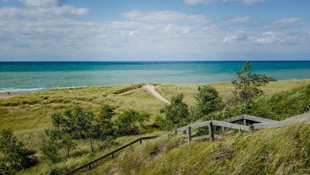 Eine Küstenlandschaft mit einem grasbewachsenen Pfad, der zu einem Strand und einer Holzkonstruktion führt, mit freiem Blick auf den Ozean.