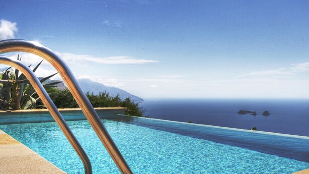 An infinity pool with clear blue water, a metal pool ladder, and a view of the ocean and distant mountains. - Villa Raffaella (Massa Lubrense)