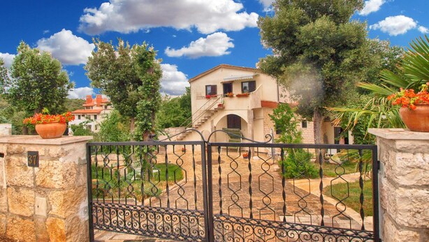 A stone gate with a metal fence leading to a building with a staircase and a balcony.