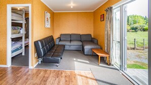 Un salon chaleureux avec un canapé d’angle, une petite table en bois et un banc intégré. Une porte vitrée donne accès à un jardin.