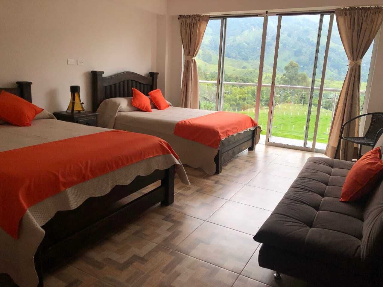 A hotel room with two beds, a sofa, and a large window offering a view of greenery.