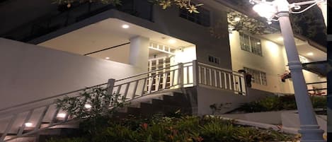 Un edificio moderno con una escalera que lleva a un balcón, rodeado de vegetación e iluminado por farolas.