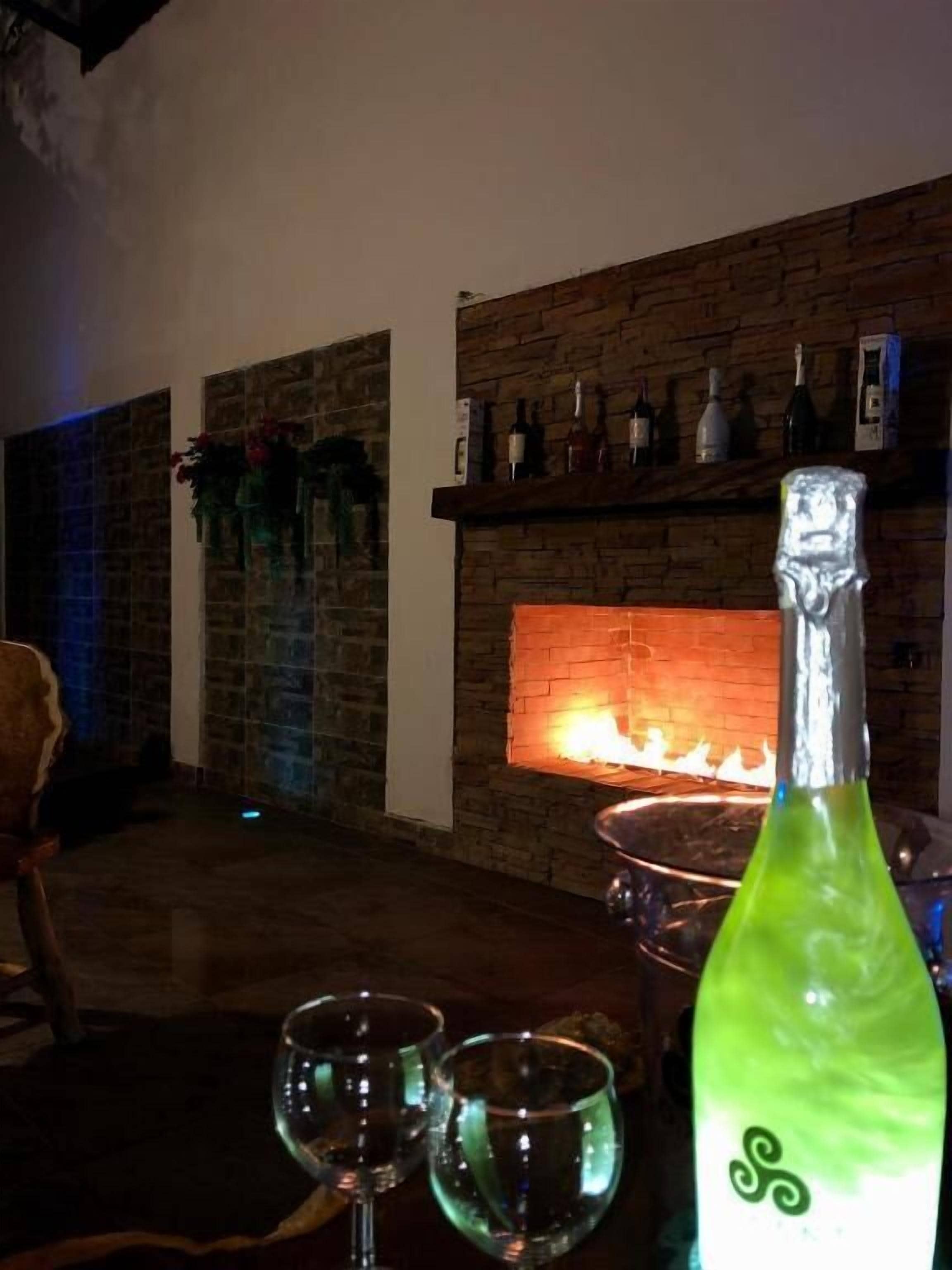 Una habitación con chimenea de ladrillo, una mesa con dos vasos y una botella, y una silla.