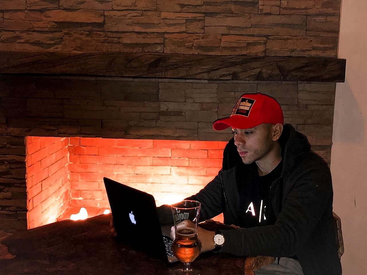 A person sitting at a table with a laptop and a glass of beer in front of a brick fireplace.