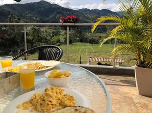 Ein Tisch mit Frühstücksangeboten und Getränken, mit Blick auf eine malerische Landschaft mit Bergen und viel Grün.