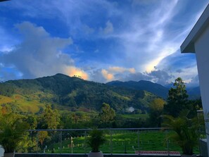 Un balcon offrant une vue sur des collines verdoyantes et une chaîne de montagnes sous un ciel partiellement nuageux.