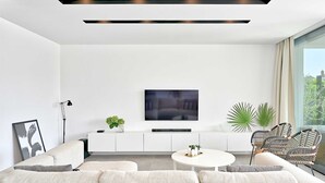 A modern living room with a white sofa, a coffee table, a TV on a white cabinet, and a large window with a view of greenery.