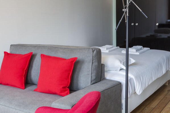 Quarto de hotel moderno com sofá cinzento, almofadas vermelhas, cama branca e abajur.