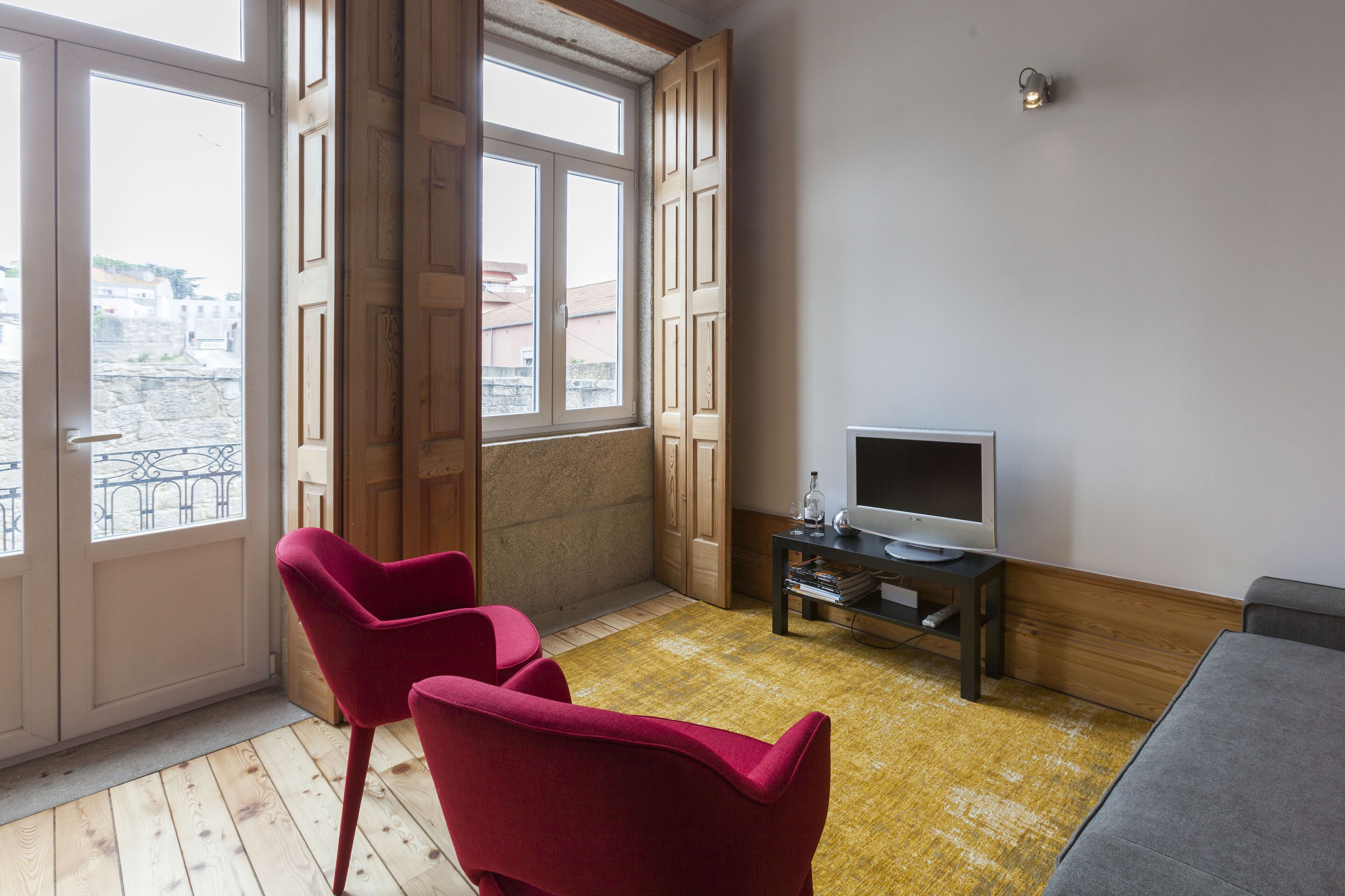 A room with a TV, two red chairs, a sofa, and large windows with a city view.