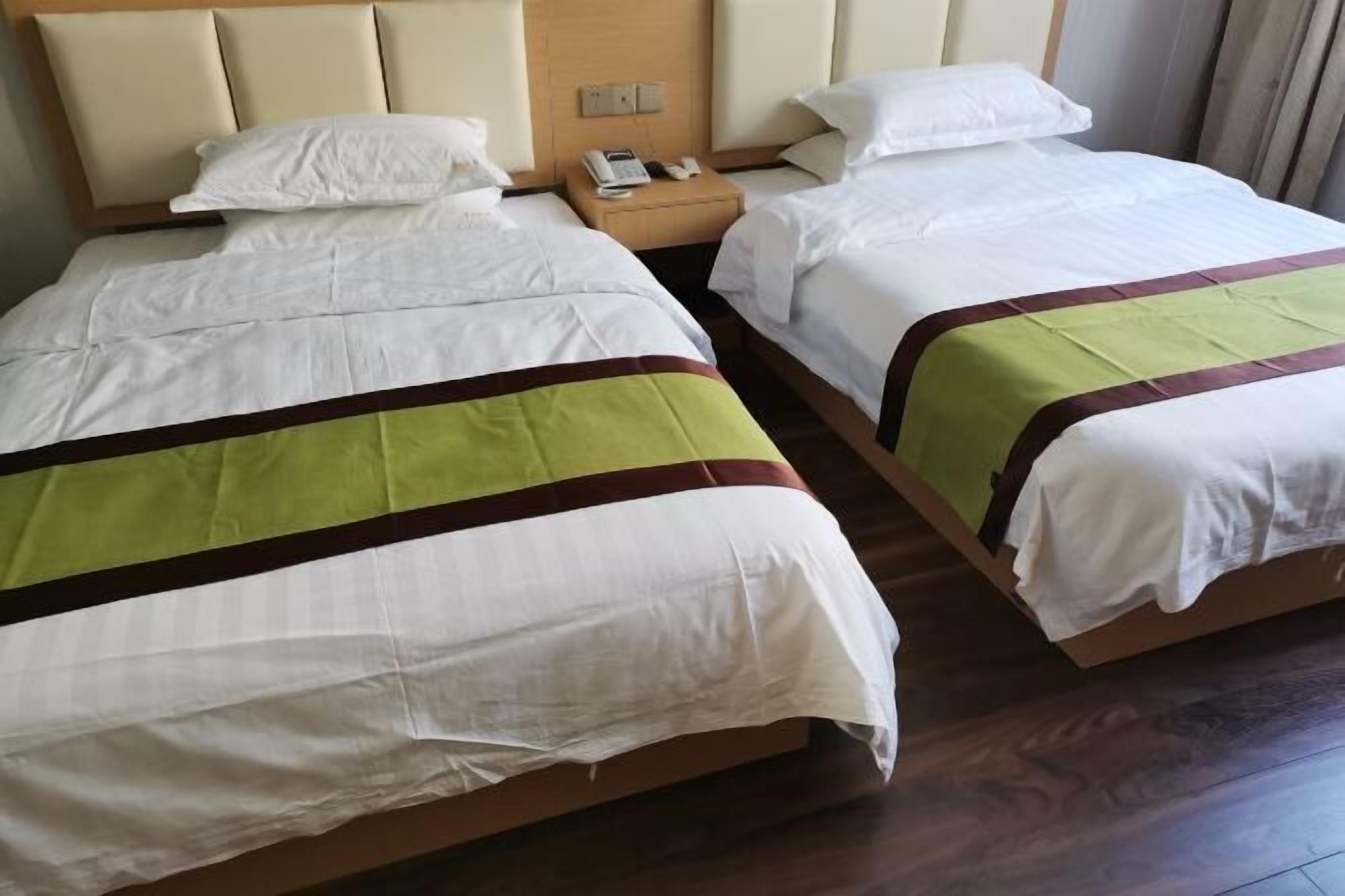 Ein Hotelzimmer mit zwei Betten, jeweils mit weißer Bettwäsche und einer grün-braunen Streifenbahn. Es gibt ein hölzernes Kopfteil, einen Nachttisch mit Telefon und Lampe sowie ein Fenster mit Vorhängen.