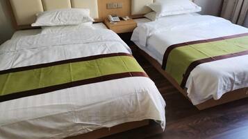 Ein Hotelzimmer mit zwei Betten, jeweils mit weißer Bettwäsche und einer grün-braunen Streifenbahn. Es gibt ein hölzernes Kopfteil, einen Nachttisch mit Telefon und Lampe sowie ein Fenster mit Vorhängen.