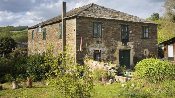 A stone building with a sloped roof, green shutters, and a small garden area in front. - A Fonte das Bodas Pensión (Samos)