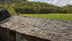 A stone-roofed building with a view of a green field and a forested hill under a partly cloudy sky. - A Fonte das Bodas Pensión (Samos)