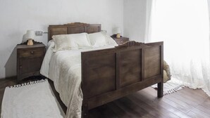 A bedroom with a wooden bed, two bedside tables with lamps, a white rug, and a window with sheer curtains. - A Fonte das Bodas Pensión (Samos)
