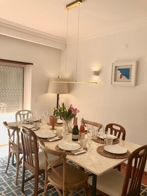 A dining area with a table set for six, featuring a white tablecloth, wooden chairs, and a vase with flowers.