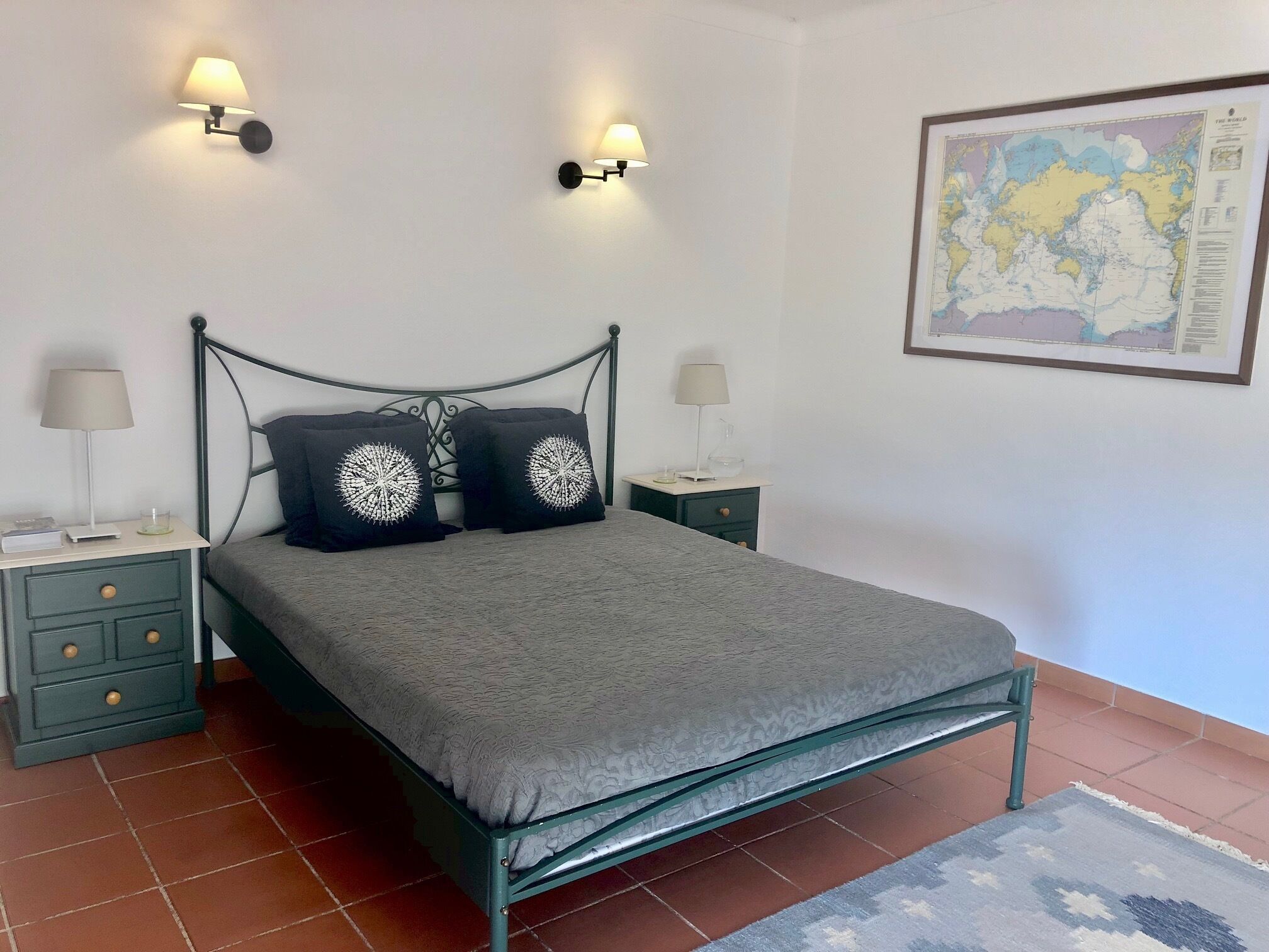 Een netjes opgemaakt slaapkamer met een metalen bedframe, twee nachtkastjes met lampen, een kaart aan de muur en een betegelde vloer.
