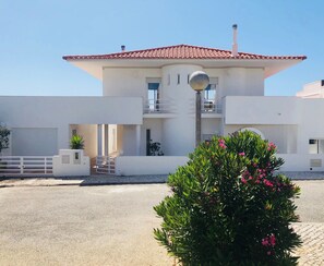 A modern two-story building with a tiled roof, a central water feature, and a small garden with pink flowers.