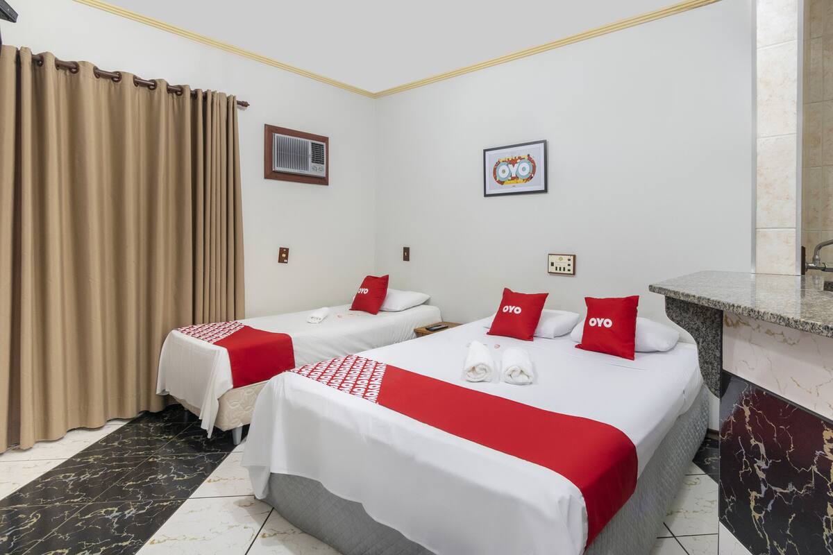 A hotel room with two beds, beige curtains, a small air conditioning unit, a framed picture, and a marble countertop.
