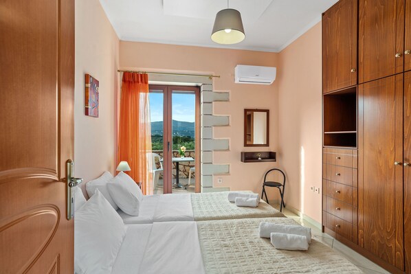 A hotel room with two beds, a wooden wardrobe, a bathroom mirror, and a view of the outdoors. - Villa Chryssa, stone villa with private pool in Neo Chorio village, Chania Crete (Neo Chorio, Crete)