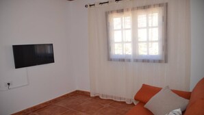 Ein Wohnzimmer mit Flachbildfernseher an der Wand, einem Fenster mit durchscheinenden Vorhängen und einem gefliesten Boden.