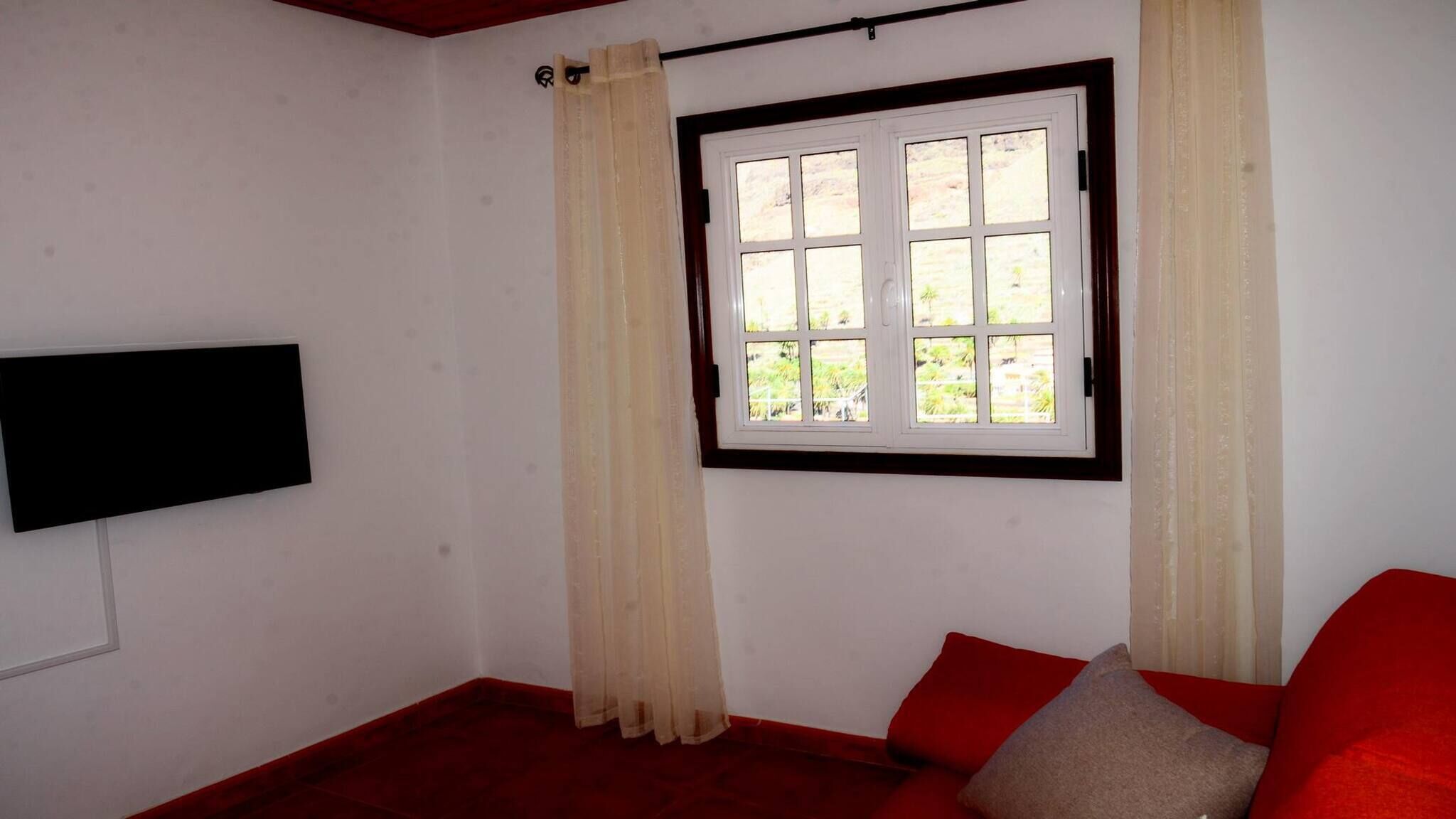 Une pièce avec une fenêtre, un canapé rouge et un téléviseur noir fixé au mur.