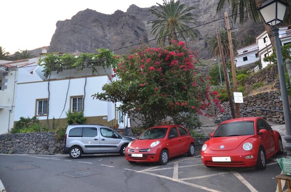 A parking area with three cars, a white building with a stone wall, and a mountain in the background. - Casita del Pedregal  (Valle Gran Rey)