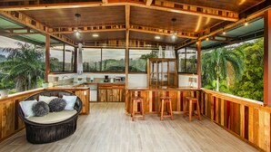 A spacious kitchen with a wooden island, bar stools, and a large window overlooking a lush landscape. - Cotingas Ocean View (Drake Bay)