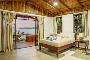A bedroom with a wooden ceiling, a bed with a white headboard, a nightstand, and a view of the sea through a sliding glass door.