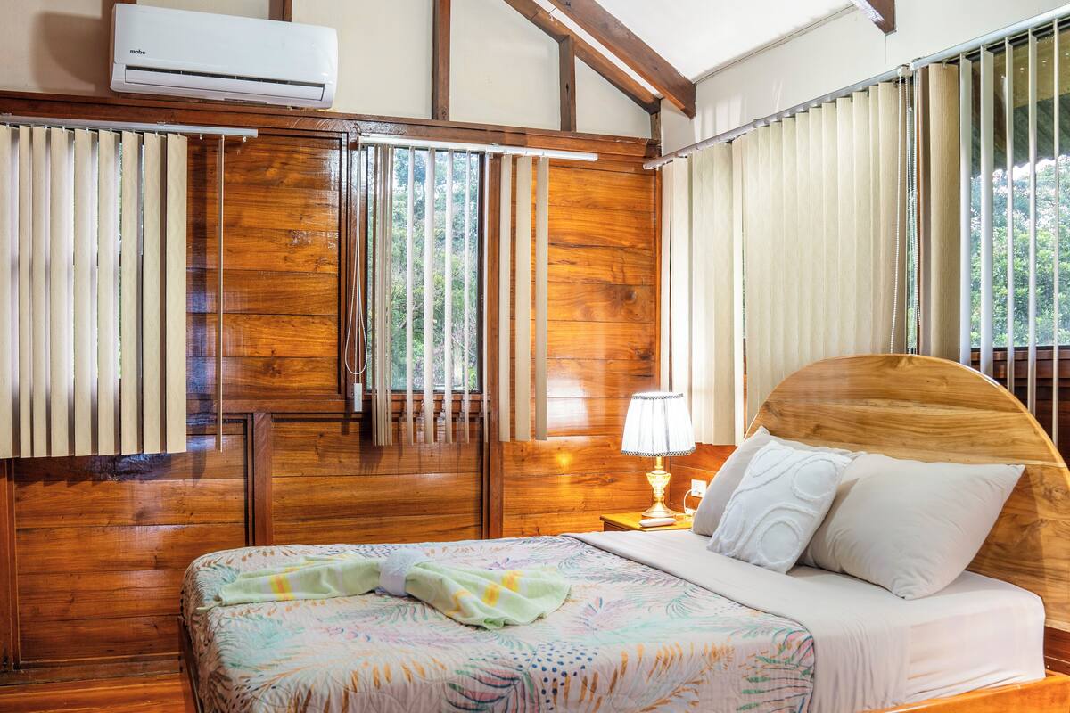 A wooden-framed bed with a floral bedspread, a bedside lamp, and a view of the outdoors through a window with vertical blinds.