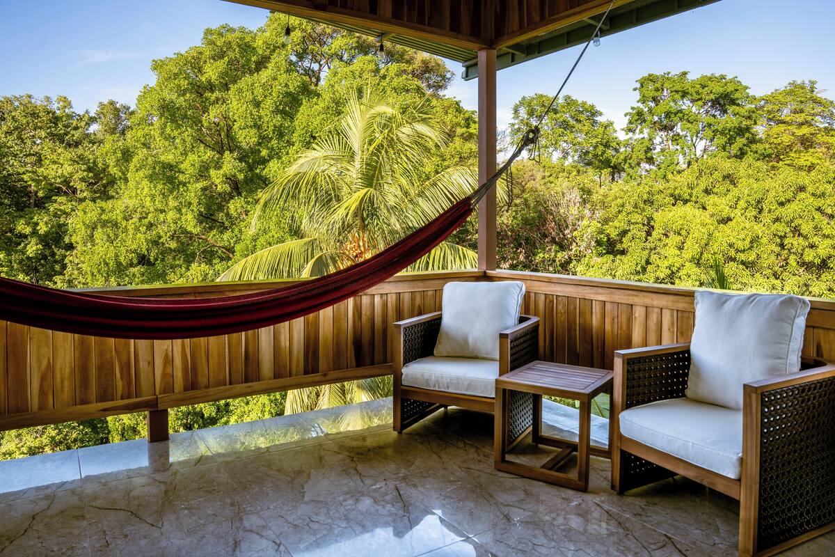 A wooden deck with two wicker chairs and a small table, overlooking a lush green forest.