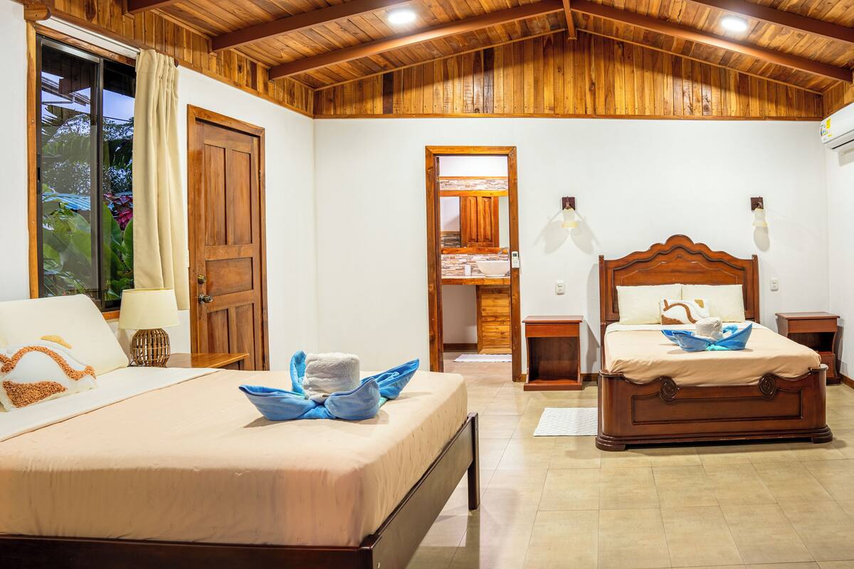 A room with two wooden beds, a wooden ceiling, a window with curtains, and a door.