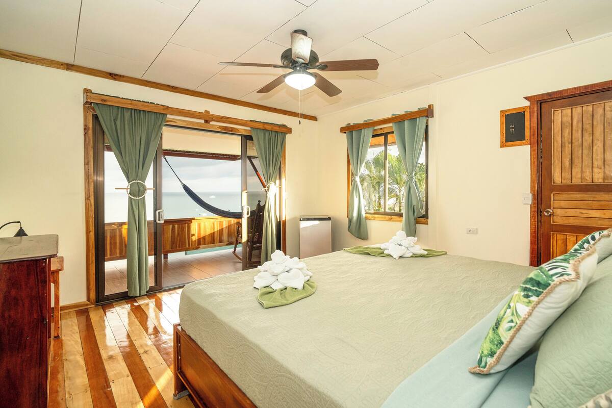 A bedroom with a bed, a desk, a ceiling fan, and a view of the ocean.