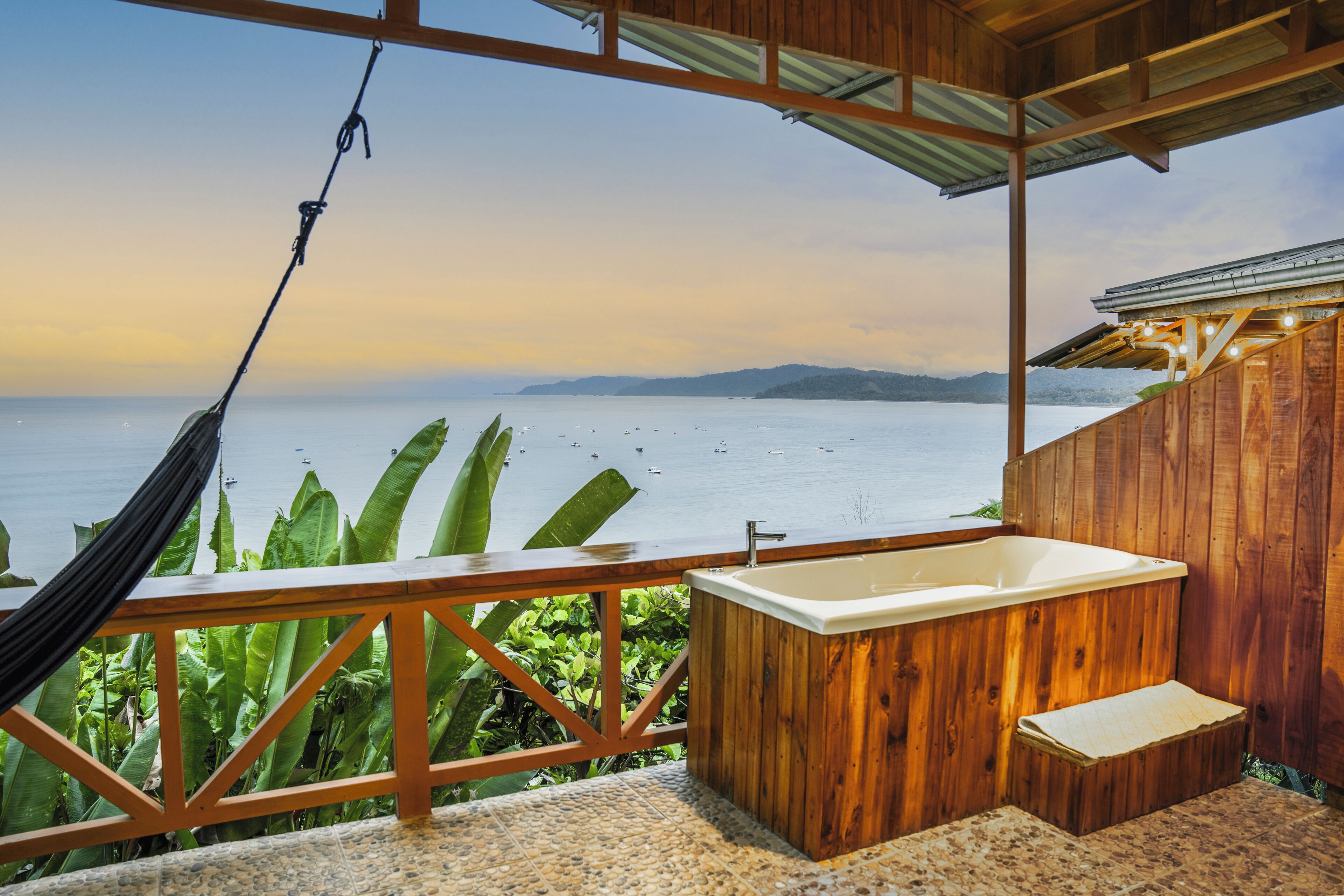 Una terraza de madera con una hamaca, una bañera de madera y vista al mar.
