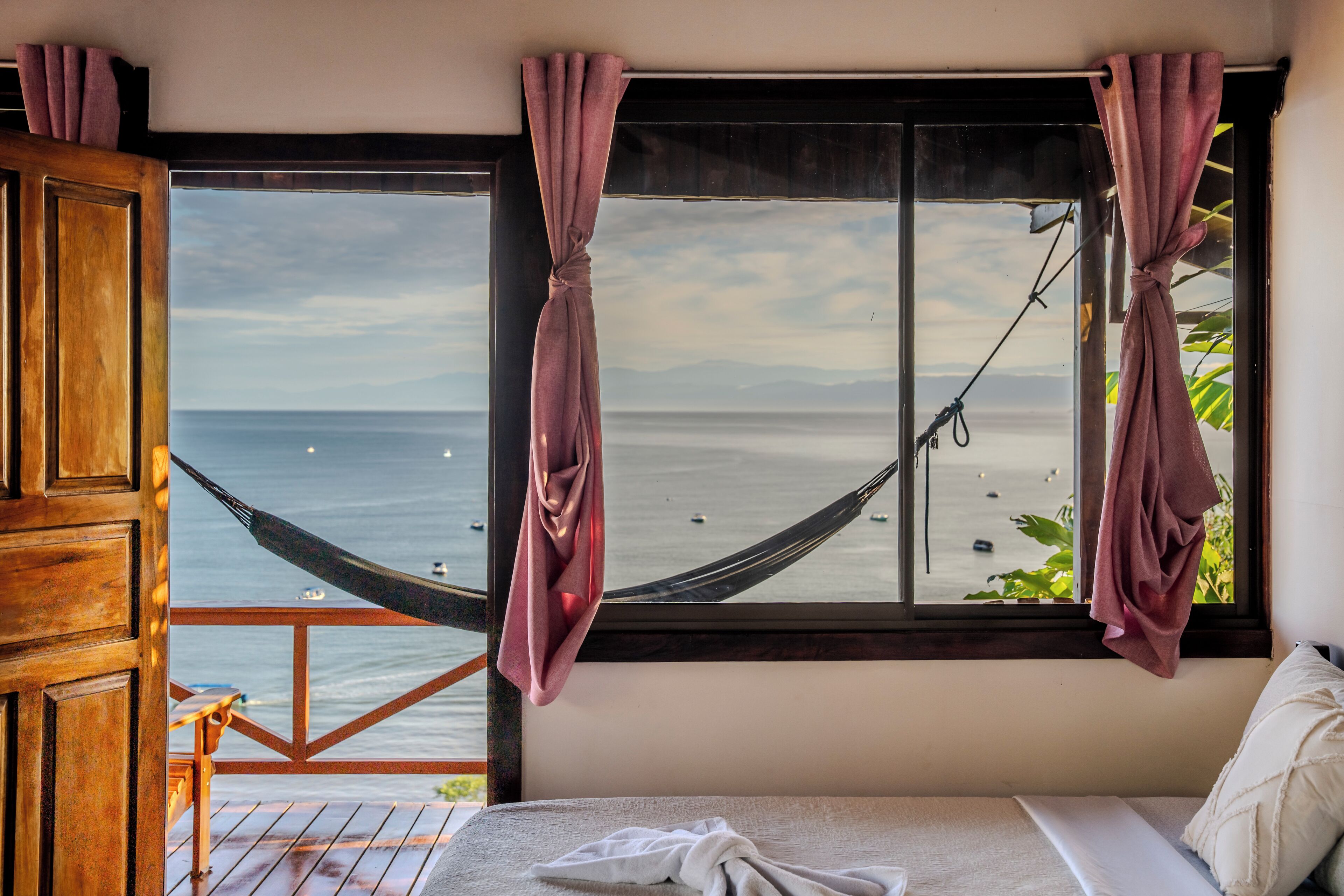 A room with a large window overlooking a beach, a hammock, and a bed with a view.