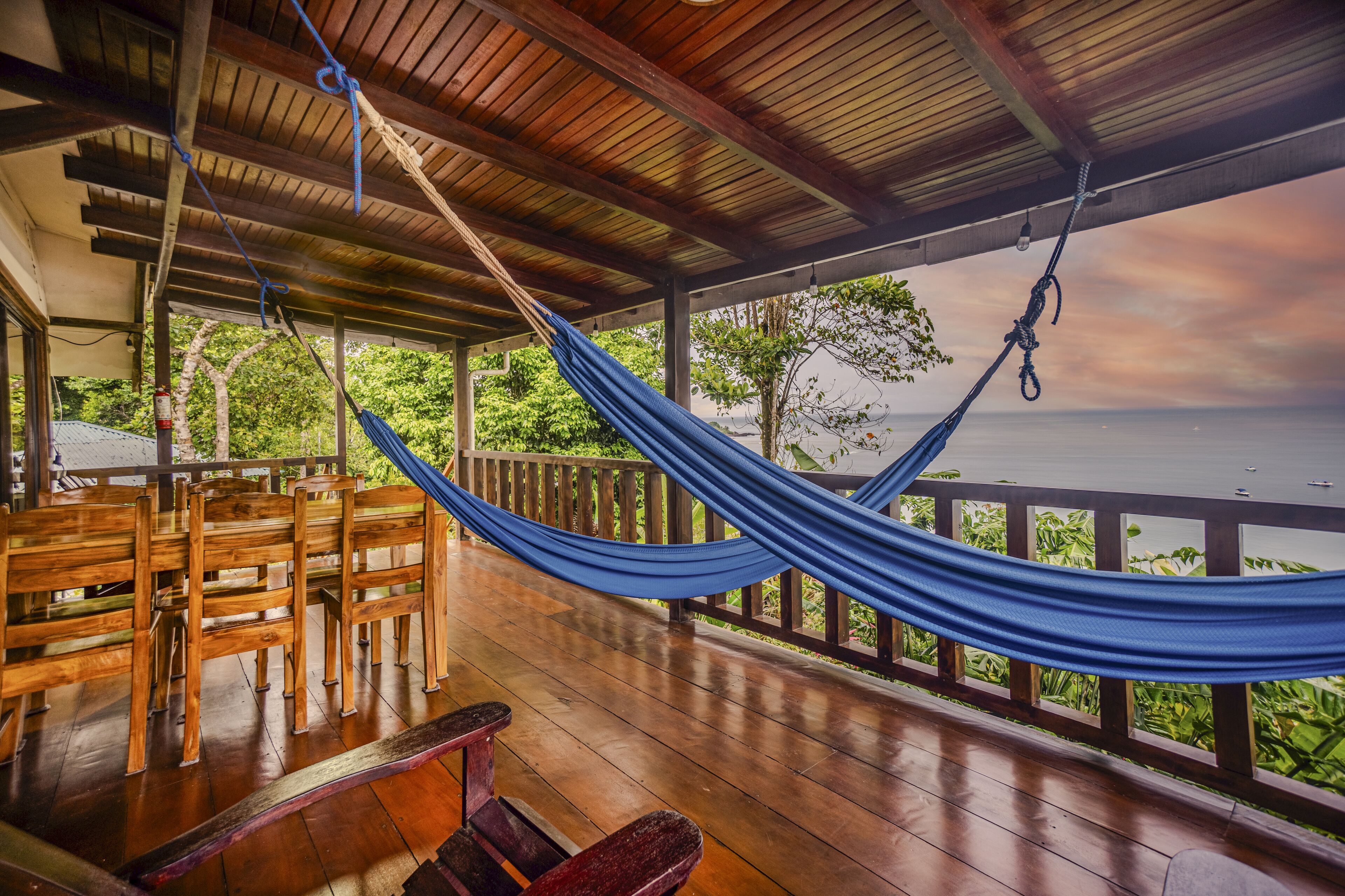 Una terraza de madera con una hamaca, sillas de madera y vista al océano.