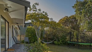 Un jardin avec une terrasse en bois, un hamac, un trampoline et une maison dotée d’un porche abrité.