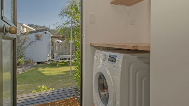Une buanderie équipée d’un lave-linge, d’une étagère en bois et d’une porte donnant sur une cour arrière avec une pelouse et un petit bâtiment.