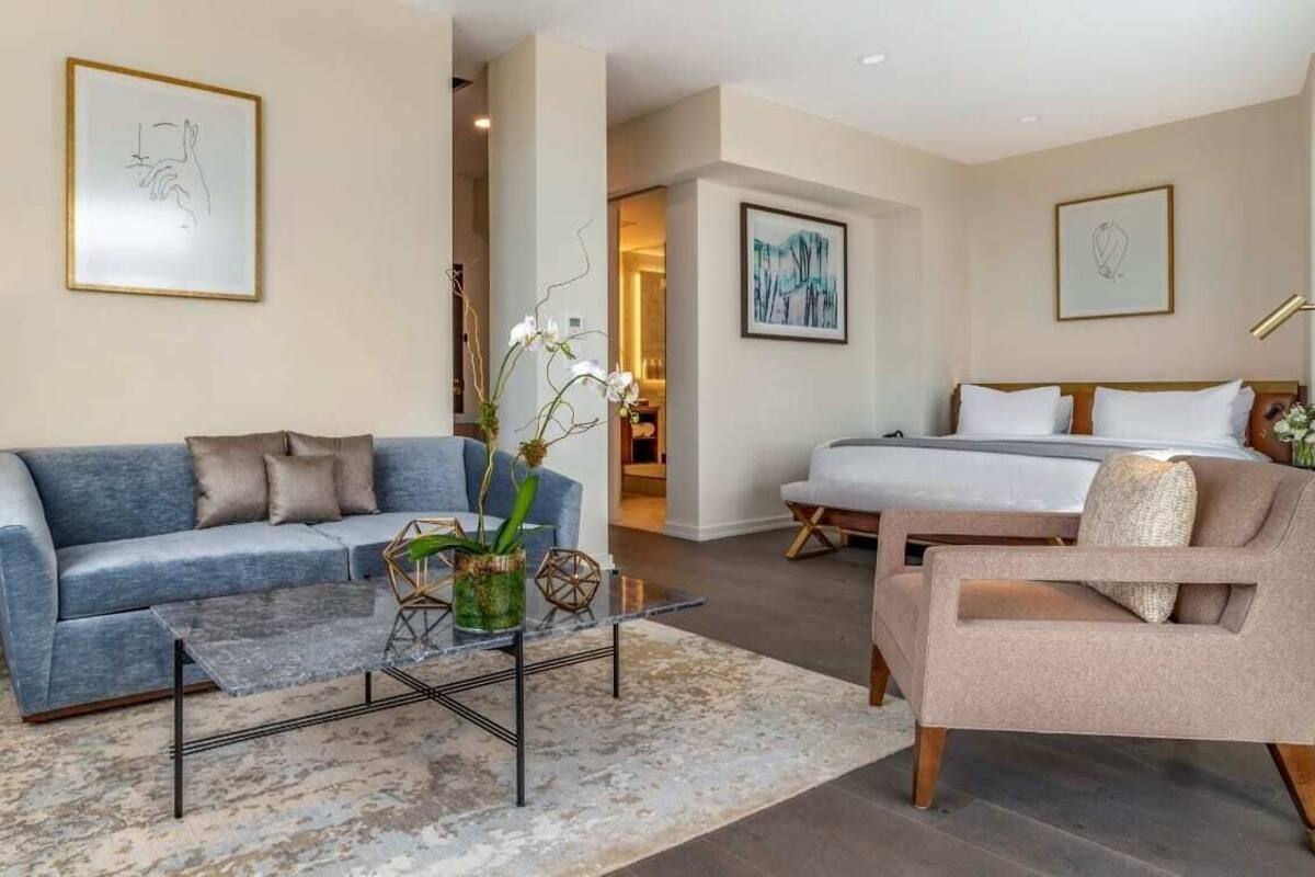 A modern hotel room with a blue sofa, a beige armchair, a bed with white linens, and a coffee table with a vase of flowers.