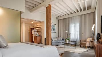 A modern hotel room with a large bed, a sofa, a coffee table, and a desk. There is a wardrobe, a mirror, and a window with curtains.
