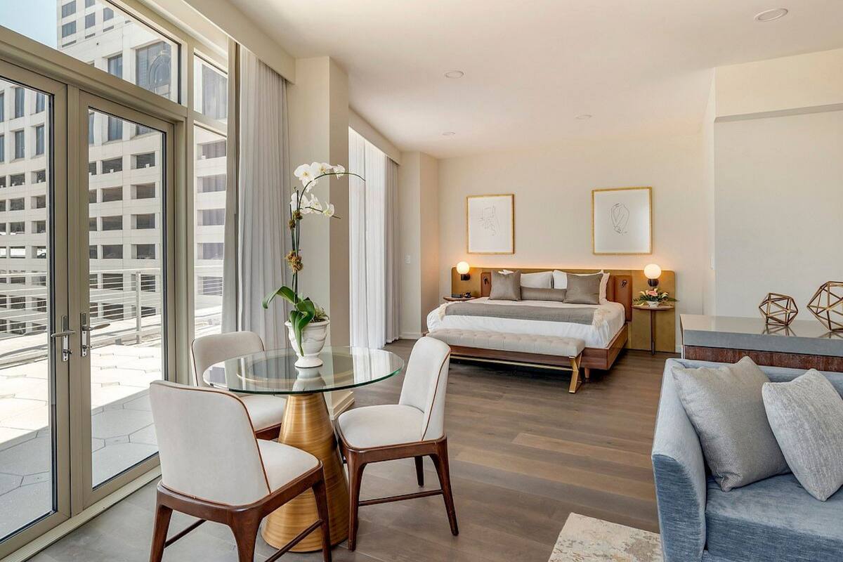 A modern hotel room with a large window, a glass dining table, a sofa, and a bed with a headboard.