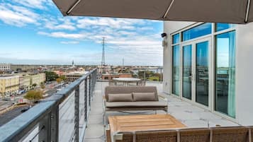 A balcony with a sofa, wooden table, and umbrella, overlooking a cityscape with buildings and a tower.