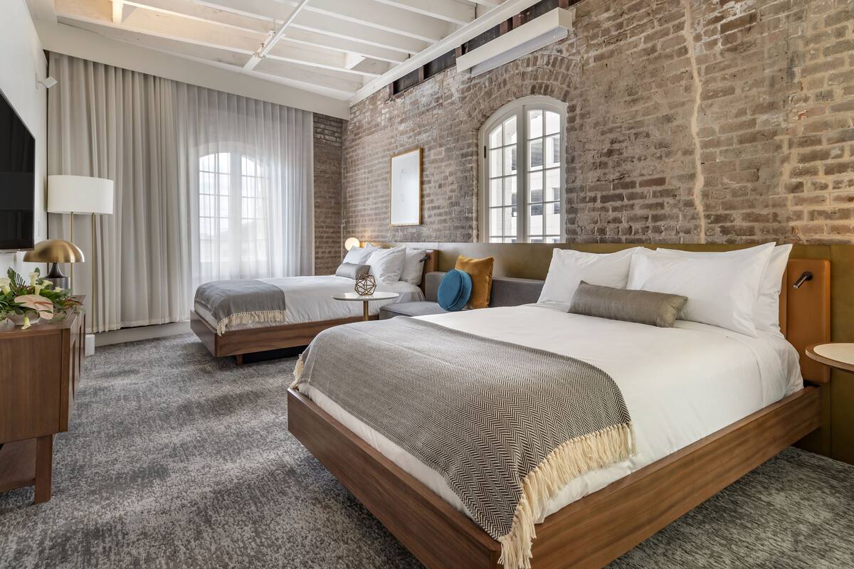 A hotel room with a large bed, a smaller bed, a brick wall, and a large window.