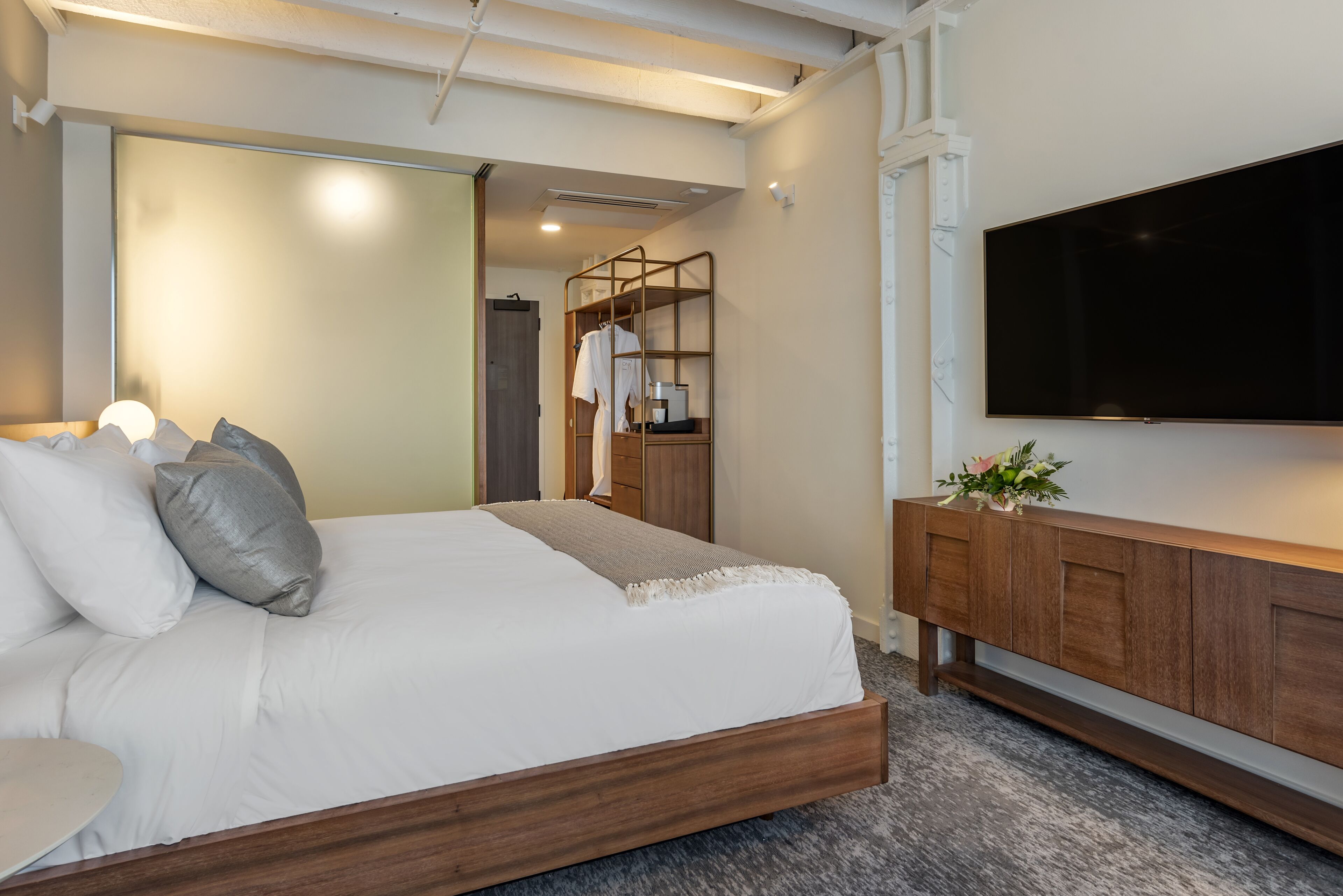 A modern bedroom with a wooden bed frame, a flat-screen TV, and a wooden console table with a vase of flowers.