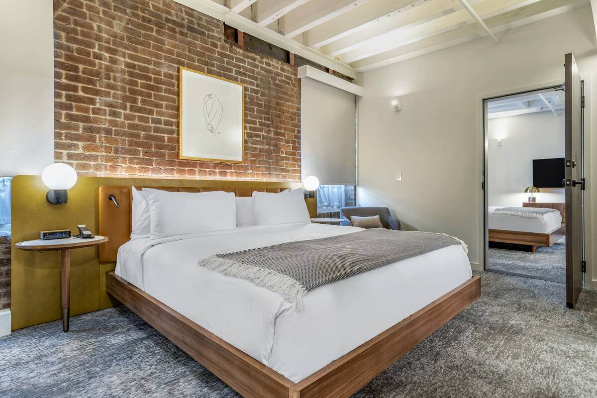A modern hotel room with a brick accent wall, a large bed with white linens, a bedside table with a lamp, and a small round table with a phone.
