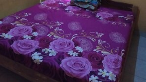 A bedroom with a bed covered in a purple floral bedspread, a window with a wooden frame, and a curtain. - Homestay Banyumili (Kalirejo)
