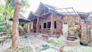 A rustic wooden building with a thatched roof, surrounded by palm trees and potted plants. - Larasati Homestay (Kalirejo)