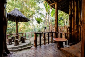 An outdoor wooden deck with rustic furniture, surrounded by lush greenery and a thatched roof structure.