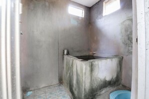 A small, dimly lit room with a concrete bathtub, a blue plastic basin, and a small window.