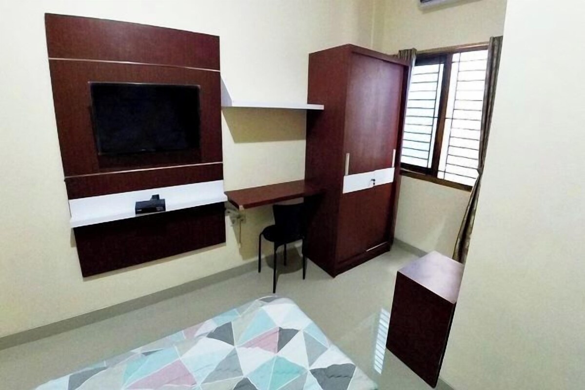 Una habitación pequeña y funcional con una televisión montada en la pared, un escritorio, una silla y una cama con un edredón estampado con motivos geométricos.