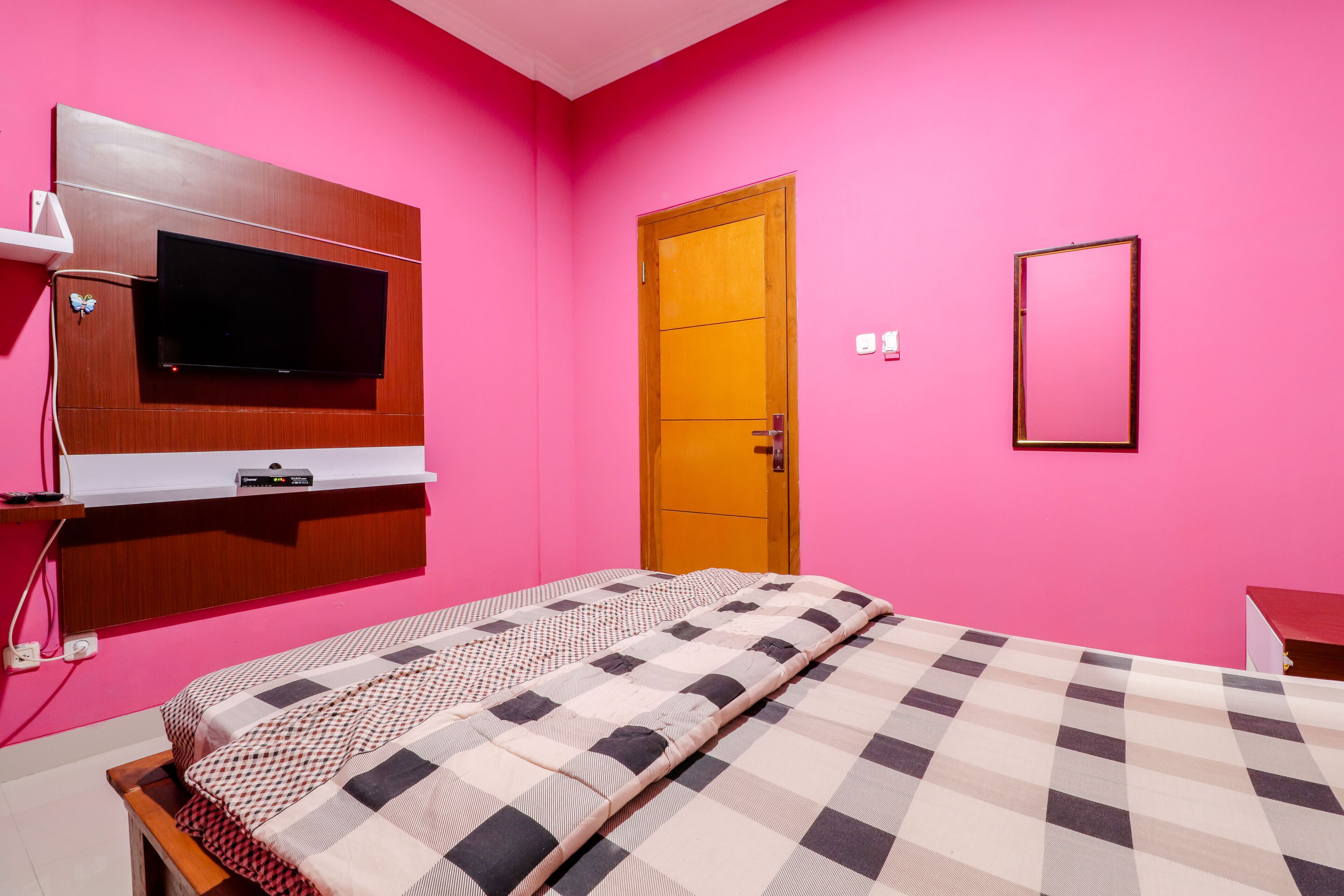 A room with a bed, a mounted TV, a wooden door, and a mirror on a pink wall.
